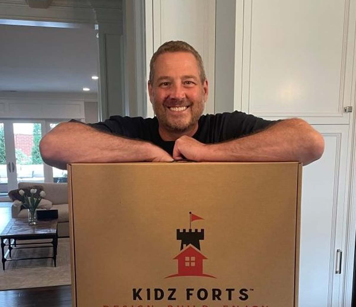 Photo of a man with a blue t-shirt smiling and posing behind a Kidz Forts cardboard carry case.