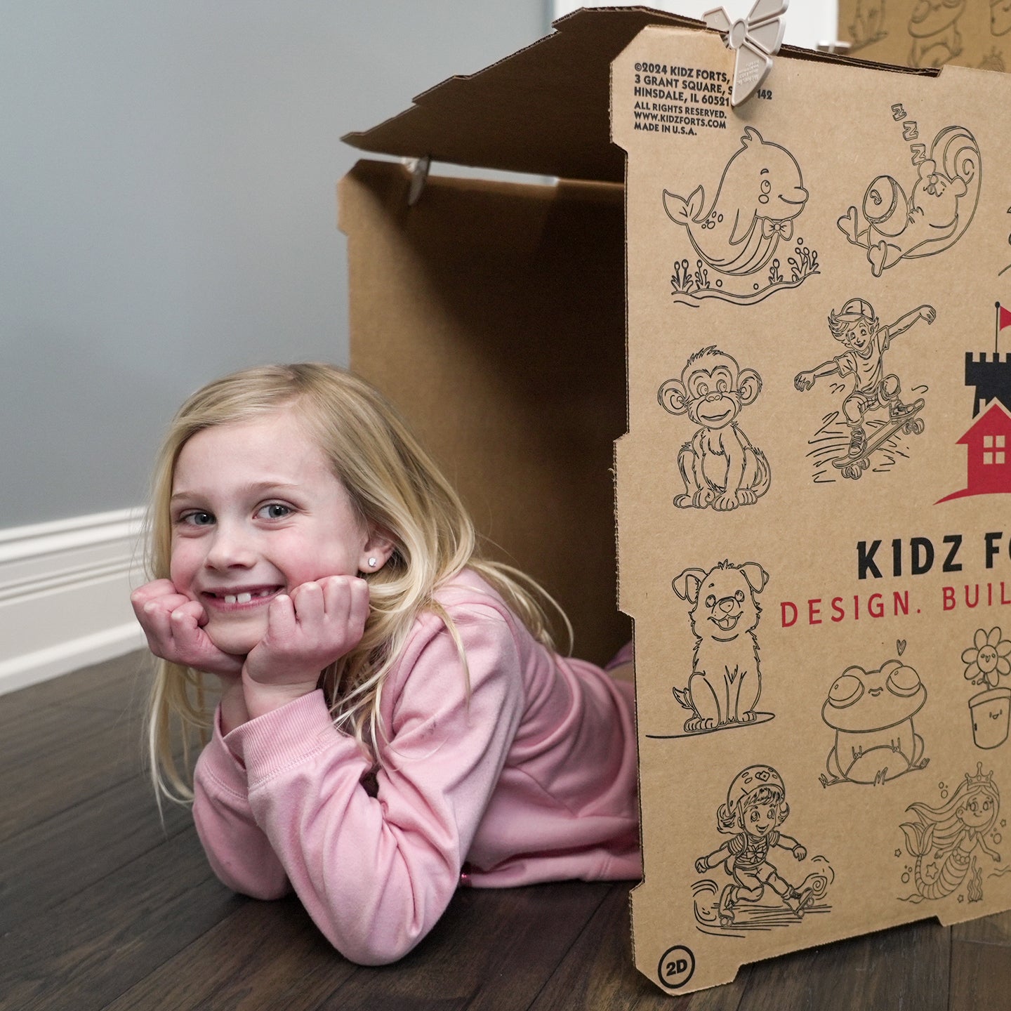 A girl in a pink sweatshirt smiling and posing on her elbows, sticking out of a Kidz Forts Hobie kit play fort.