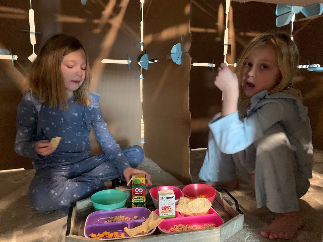 Two elementary school aged kids having lunch together and playing inside a cardboard fort.