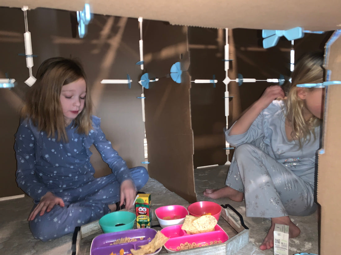 Two 3rd grade girls inside a cardboard play fort having lunch together.