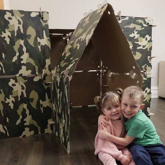 Two pre-schoolers hugging and sitting in front of a camouflage cardboard play fort.