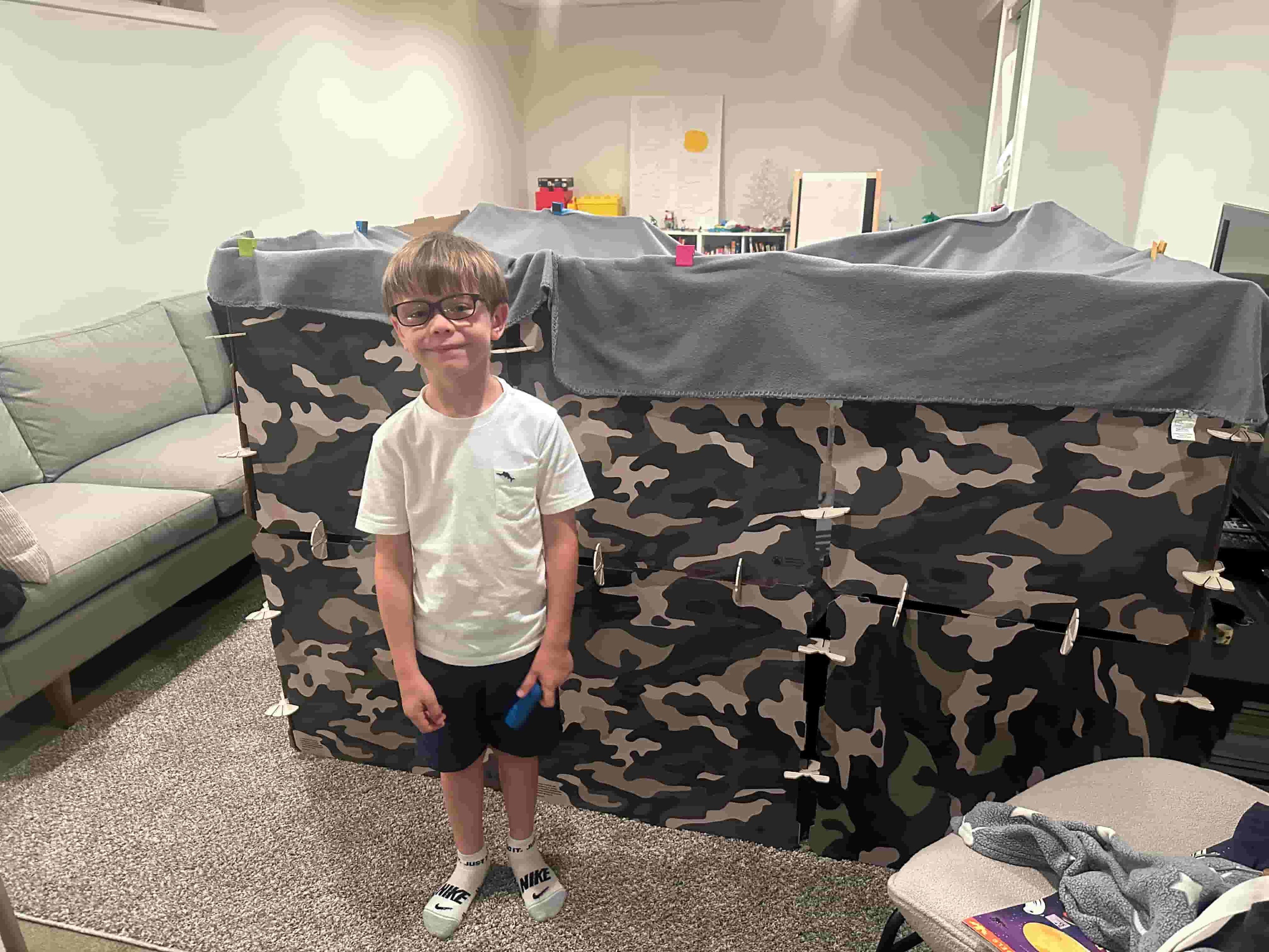 Child standing in front of a cardboard play fort printed in a camouflage pattern.