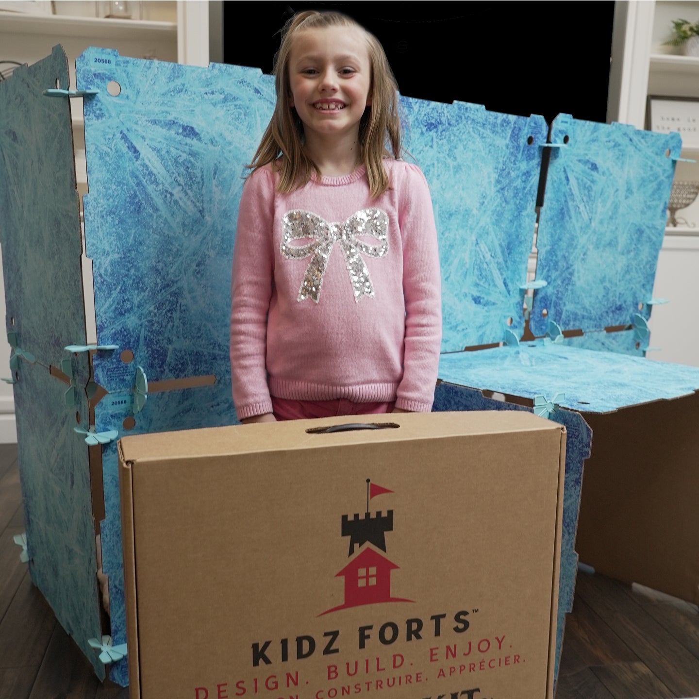 A photo of a girl in a pink sweatshirt smiling and posing with a Kidz Forts Frozen kit play fort behind her.  She's standing behind the cardboard carry case for the kit.