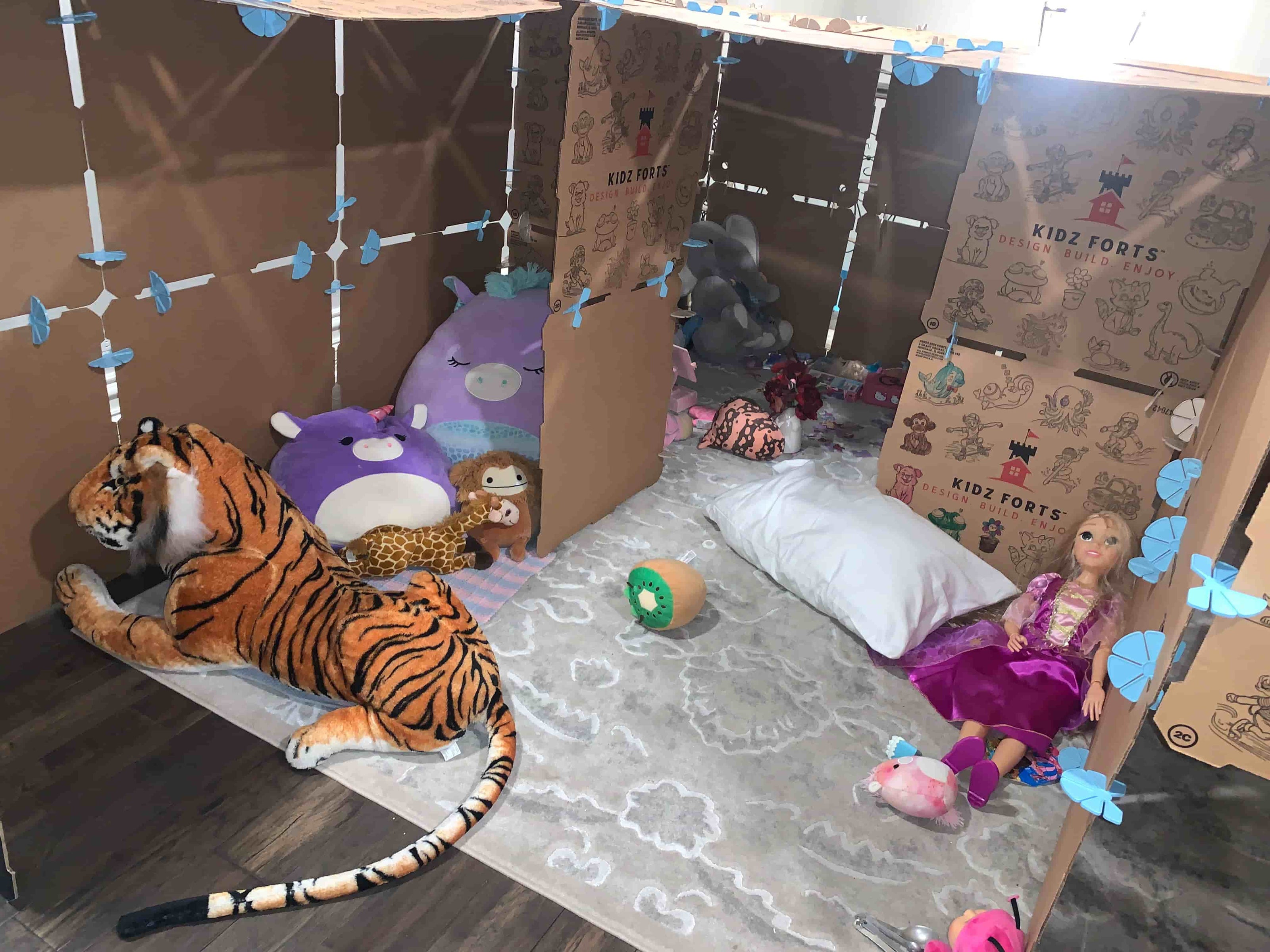 Cardboard play fort by Kidz Forts with plush toys including a tiger, purple creature, and dolls on a wooden floor.