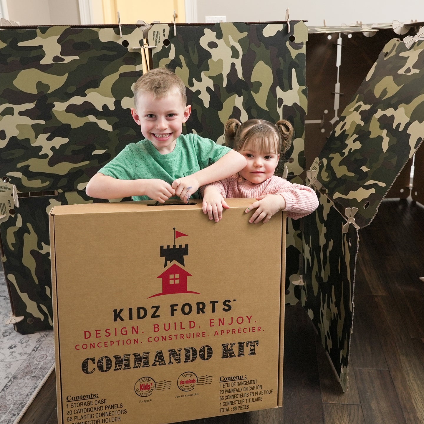 A boy and girl smiling and standing behind a Kidz Forts Commando Kit carry case.  Camouflage children's play fort in the background.